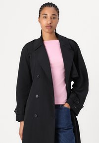 Cappotto trench nero doppiopetto con grandi bottoni, ampi revers e una vestibilità rilassata. Indossato sopra un top rosa, abbinato a jeans blu.