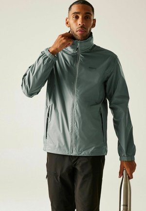 Hombre ajustándose el cuello de una chaqueta ligera gris, sosteniendo una botella de agua de acero inoxidable, de pie frente a un fondo liso.