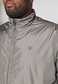 Man wearing a gray zip-up jacket with a high collar featuring red, white, and navy stripes and a small rectangular logo on the chest.