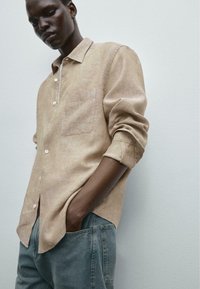 Linen button-up shirt in light beige with a chest pocket and rolled sleeves, paired with blue denim pants. Smooth texture and relaxed fit.