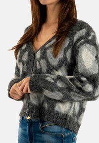 Grijze en witte fluffy cardigan met een V-hals, knoopsluiting en een luipaardprint. Materiaal lijkt zacht en gestructureerd.
