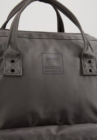 Sac en tissu texturé gris foncé avec double poignée, coutures renforcées, rivets noirs et un logo carré discret à l'avant.