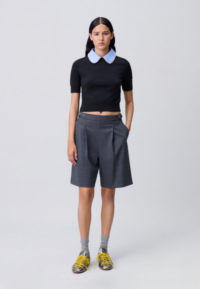 Top negro de manga corta con un cuello en contraste de color azul claro, combinado con pantalones cortos grises con pliegues. Completado con calcetines grises y zapatillas con detalles amarillos.