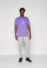 Camiseta de algodón morada con diseño gráfico geométrico en el pecho, combinada con pantalones deportivos gris claro con franjas laterales blancas y zapatillas oscuras.