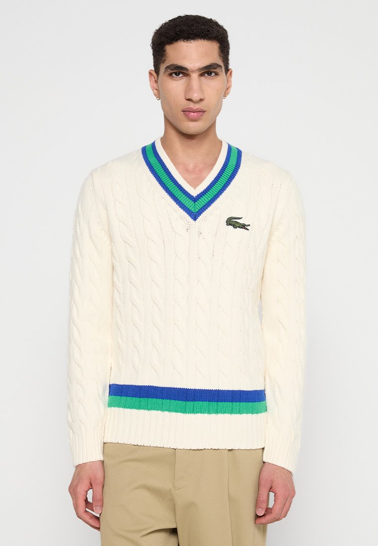 Uomo che indossa un maglione beige a trecce con scollo a V, strisce blu e verdi e logo coccodrillo ricamato, abbinato a pantaloni beige.