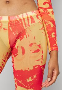 Leggings feitos de um tecido elástico com um padrão abstrato vibrante em vermelho e amarelo, apresentando elementos gráficos e uma textura suave.