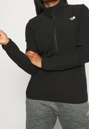 Osoba nosząca czarną zapinaną na zamek kurtkę North Face z długimi rękawami oraz szare teksturowane spodnie, z rękami skrzyżowanymi na wysokości talii.
