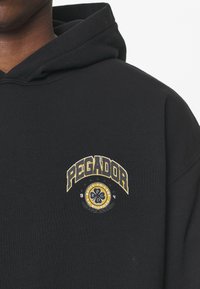 Felpa nera con cappuccio in cotone, con logo "Pegador" giallo e bianco caratterizzato da accenti circolari e testo decorativo.