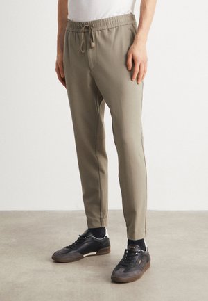 Mand iført beige slim-fit joggebukser med elastisk talje og sorte lave sneakers på en lys gulv og hvid baggrund.