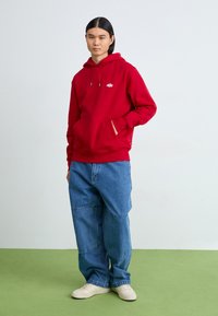 Hanorac roșu cu buzunar frontal și glugă cu șnur, purtat împreună cu pantaloni largi din denim albastru și adidași albi, pe o suprafață verde.