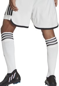 Weiße Sportshorts mit schwarzem Saum, kombiniert mit weißen Socken mit drei schwarzen Streifen. Schwarze Fußballschuhe mit blauen Akzenten.