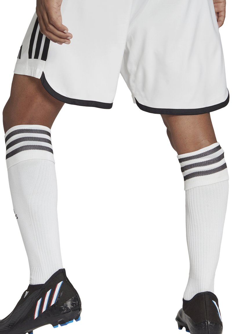 Weiße Sportshorts mit schwarzem Saum, kombiniert mit weißen Socken mit drei schwarzen Streifen. Schwarze Fußballschuhe mit blauen Akzenten.