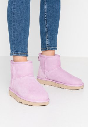 Henkilö, joka käyttää vaaleanvioletteja mokkanahkaisia nilkkavarsikenkiä, joissa on beiget pohjat, ja sinisiä kapeita farkkuja, seisomassa valkoisella lattialla.