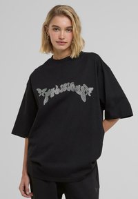 Maglietta nera oversized in cotone con maniche corte e scollatura rotonda, caratterizzata da un logo decorato con strass.