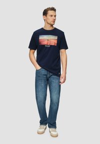 Marineblauwe katoenen T-shirt met een rechthoekige grafische print van een auto op een onverharde weg. Gecombineerd met lichtblauwe spijkerbroeken en sneakers.