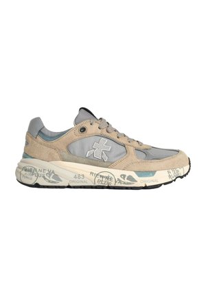 Zapatilla beige y gris con cordones y suela gruesa que muestra texto impreso y logotipos, parte superior de ante y malla, y un emblema cruzado estilizado en el lateral.