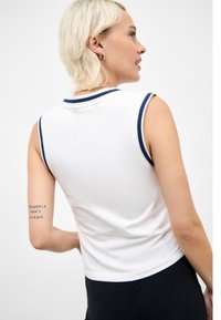 White sleeveless top with navy accents on the collar and armholes. Smooth texture with a fitted design. Black pants partially visible at the bottom.