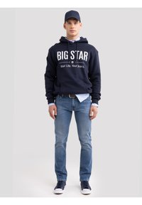 Mees, kellel on seljas mereväe toonides kapuutsiga dressipluus tekstiga "BIG STAR Your Life. Your Jeans.", helesinised teksapüksid, mereväe värvi nokamüts ja mereväe toonides tennised, seisab valge tausta ees.