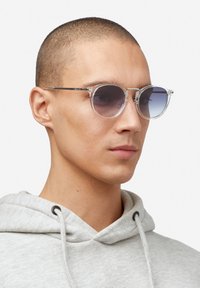 Prozirne okrugle sunčane naočale s tamnim gradijentnim staklima, prozirnim okvirom i tankim srebrnim metalnim detaljima. Naočale nosi osoba u sivom kapuljaču.