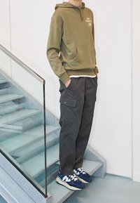 Olivgrön hoodie med framficka och logotyp, ihop med löst sittande mörkgrå cargobyxor med sidofickor och marinblå sneakers.