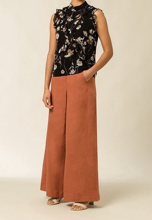 Vrouw draagt mouwloze zwarte blouse met bloemenruche, wijde roestkleurige broek en beige open-teen sandalen met hak, staat met één hand in de zak.