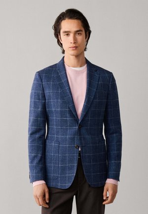 Jeune homme portant un blazer bleu à carreaux par-dessus un pull rose et un pantalon noir, debout devant un fond clair uni.