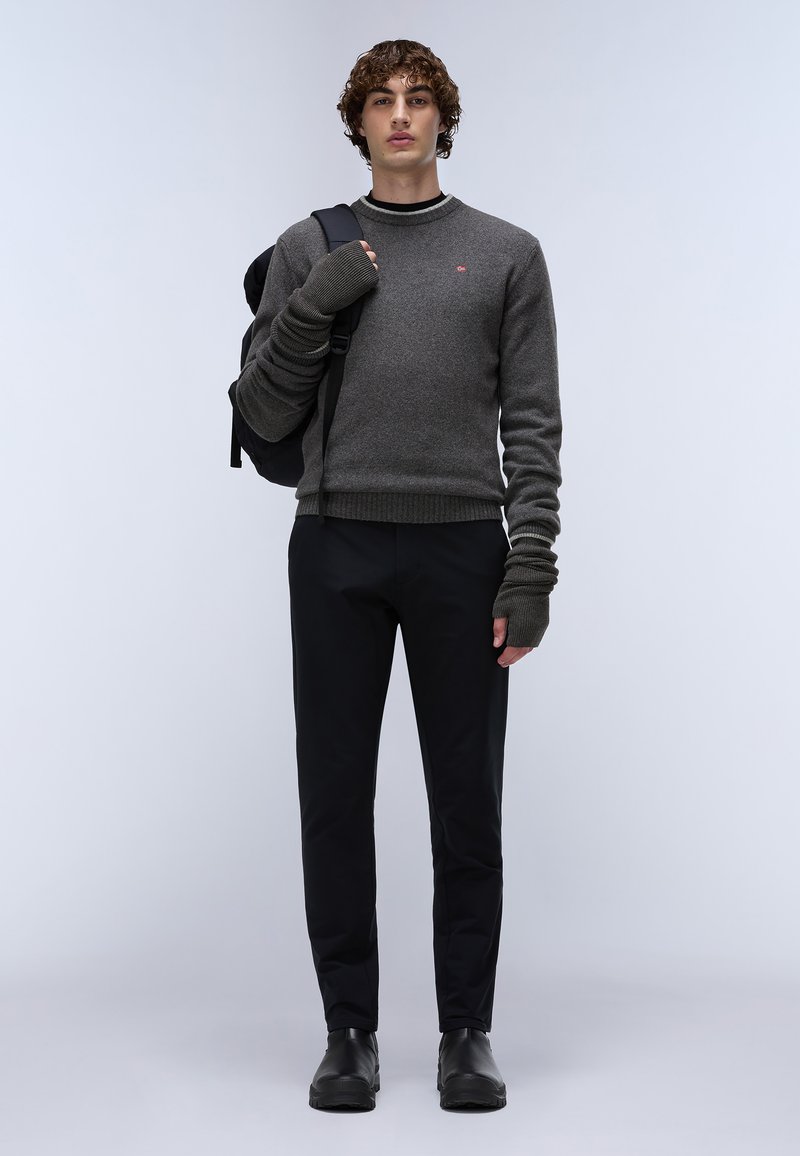 Szary sweter z dzianiny z długimi, prążkowanymi rękawami, w połączeniu z czarnymi spodniami slim fit i czarnymi butami. Modelka trzyma czarny plecak.