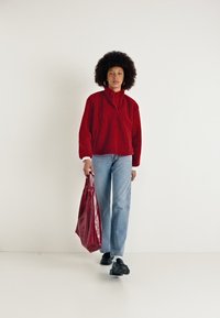 Rød fleecegenser med høy krage, kombinert med lys blå rett passform jeans. Holder en blank rød veske og bruker svarte sneakers.