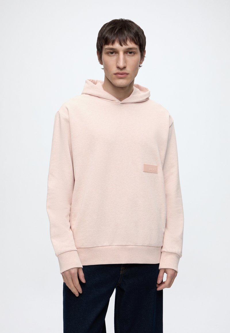 Jeune homme portant un sweat à capuche rose pâle avec un petit patch "BOSS" sur la poitrine, associé à un jean bleu foncé, sur un fond clair uni.