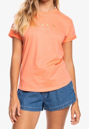 Kvinde iført en koral-farvet t-shirt med korte ærmer med "ROXY Since 1990" i gyldent tryk og blå denimshorts.