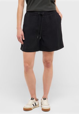 Person wearing black drawstring shorts, black top, and white sneakers with black stripes, standing against a plain light background.