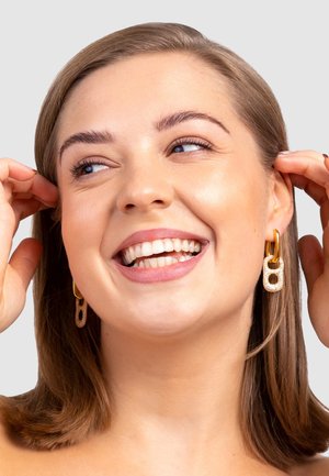 Gold- und Silberohringe mit einem Kettenglieddesign, das strukturierte Elemente und glatte Akzente aufweist, getragen von einer Person mit schulterlangen Haaren.