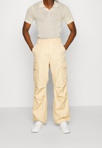 Kremowy sweter polo z krótkimi rękawami, zestawiony z jasnobeżowymi spodniami cargo z dużymi bocznymi kieszeniami. Całość dopełniają białe tenisówki.