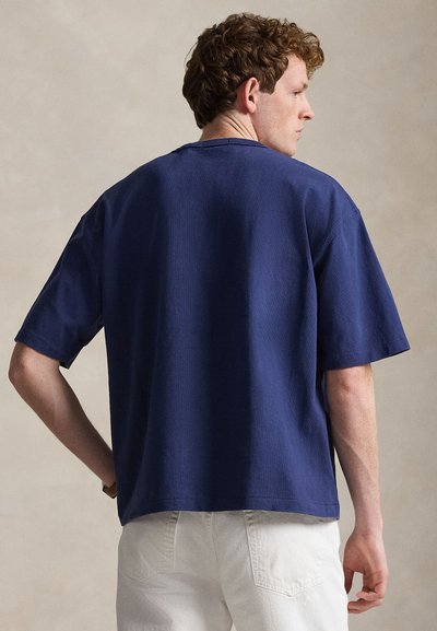 Άνδρας με σγουρά μαλλιά που φοράει μια άνετη μπλε σκούρα κοντομάνικη μπλούζα και λευκό παντελόνι, γυρισμένος πλάτη σε απλό φόντο.