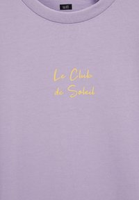 Tricou lavandă cu guler rotund și text galben „Le Club de Soleil” centrat pe piept și o mică etichetă de marcă neagră în interiorul gulerului.