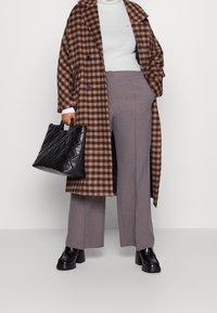 Manteau à carreaux marron et noir, col roulé blanc, pantalon large gris, sac fourre-tout matelassé noir et chaussures noires imposantes.