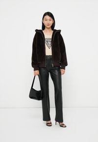 Veste en fausse fourrure marron avec capuche, T-shirt graphique crème, pants en fausse cuir noir et sac à main noir. Le modèle porte des talons noirs à lanières.