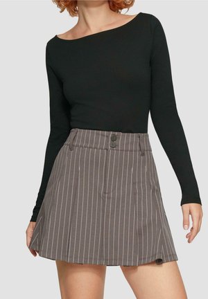 Kvinde iført en sort bluse med lange ærmer og en grå plisseret nederdel med nålestriber og to knapper i taljen mod en ensfarvet baggrund.