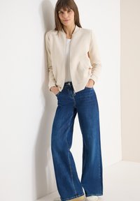 Femme aux cheveux bruns portant une veste crème zippée, un haut blanc, un jean bleu à jambes larges, des baskets blanches, debout contre un mur blanc les mains dans les poches.