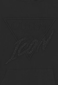 Schwarzer Stoff mit geprägt Guess-Logo und der Schrift "ICON", verziert mit kleinen schwarzen Perlen.