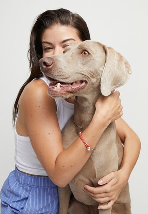 Jeune femme en débardeur blanc et pantalon bleu, serrant dans ses bras un grand chien marron clair avec un collier rouge, sur un fond blanc uni.