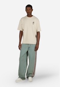 Bež majica od pamuka s kratkim rukavima i malim grafičkim printom, u kombinaciji s širokim svijetloplavim trapericama s bočnim trakama.