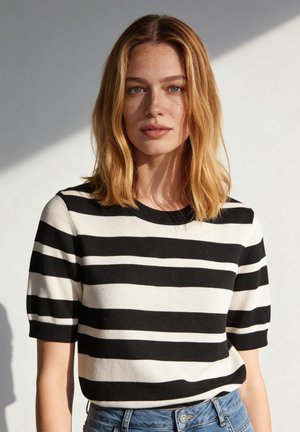 Młoda kobieta o blond włosach do ramion, ubrana w czarno-biały pasiasty krótki top i niebieskie dżinsy, stoi przy białej ścianie.