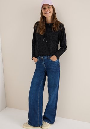 Svart leopardmönstrad sweatshirt med snörning i huvan, kombinerad med vida blå jeans och beige sneakers, bärs med en ljuslila keps.