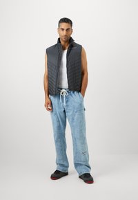 Párnázott fekete mellény egy fehér trikó felett, laza világoskék farmernadrággal párosítva. A nadrágon Nike logó található, és fekete sportcipők vannak rajta.