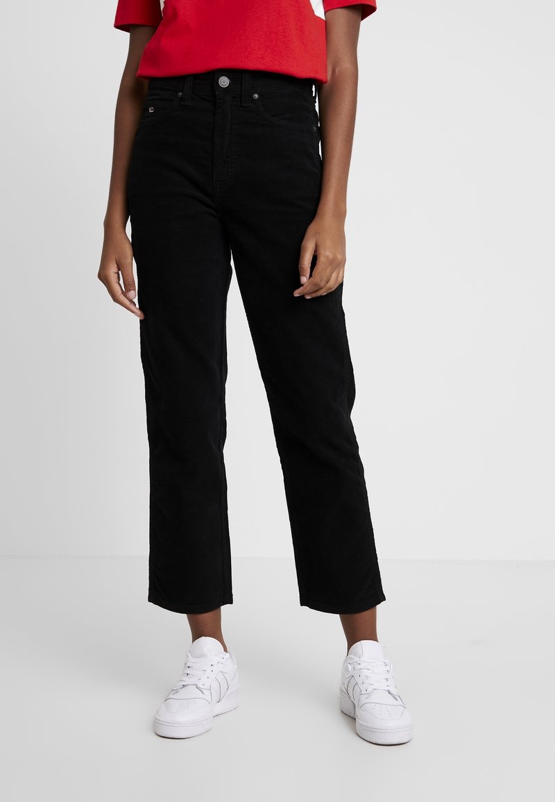 Pantalones negros de pana con un diseño de pierna recta, cintura alta y textura sutil, combinados con zapatillas blancas y una camiseta roja.