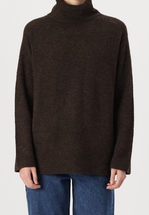 Osoba ubrana w ciemnobrązowy prążkowany sweter z golfem i długimi rękawami oraz niebieskie dżinsy na jednolitym tle.