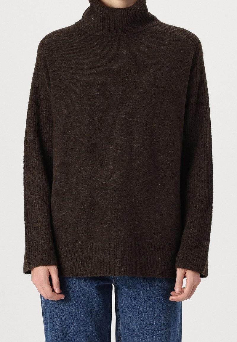 Personne portant un pull à col roulé côtelé brun foncé à manches longues et un jean bleu en denim, sur un fond uni.