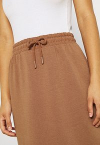 Pantalon de survêtement marron avec une taille à cordon, fabriqué en tissu doux. Présente des coutures visibles et un design fuselé.