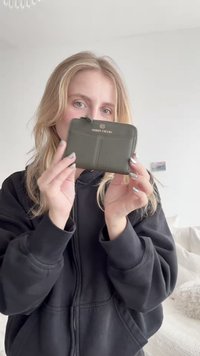 Portfel z oliwkowej zielonej skóry o fakturowanym wykończeniu, zamykany na zamek błyskawiczny, z ozdobnym logo z przodu. Mały i prostokątny kształt.